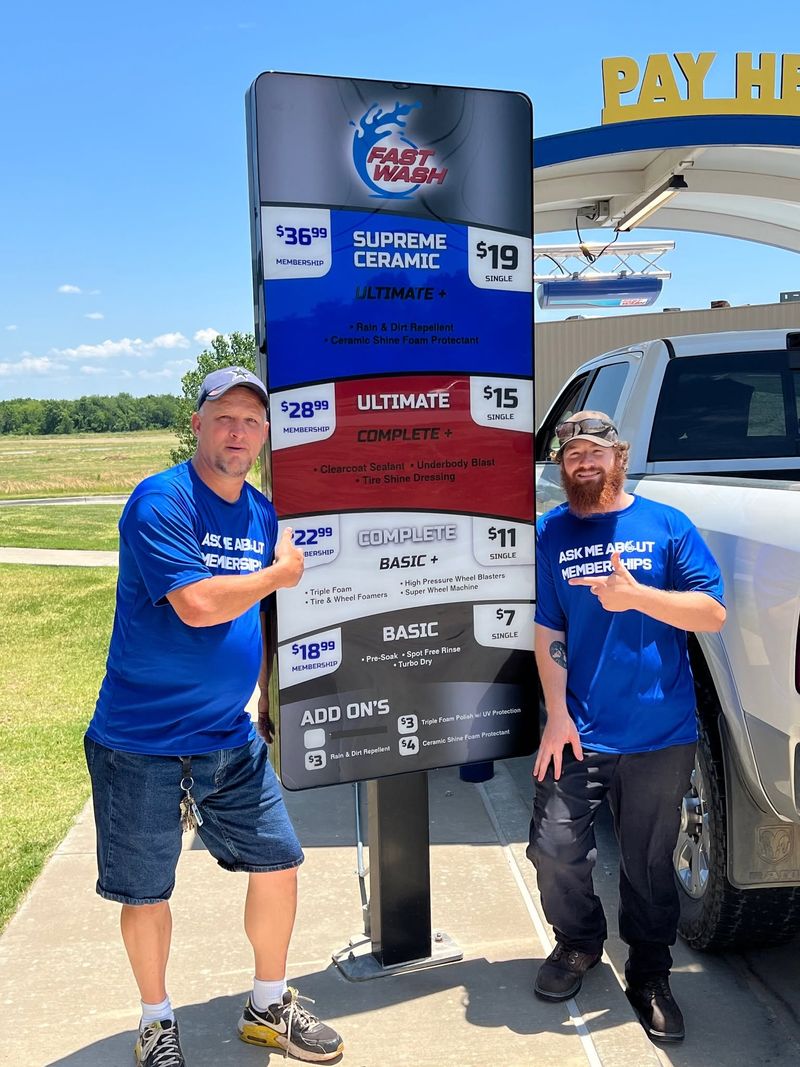Two Fast Wash crew members in blue Ask Me About Memberships shirts pointing at the wash menu board by the pay station