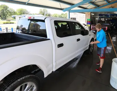 Crew member pressure-rinsing the side of a white pickup at the entrance, before the tunnel