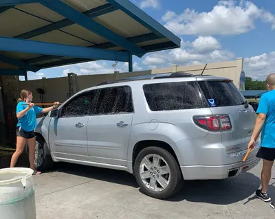 Two crew members in blue shirts scrubbing bugs off a silver SUV before it enters the tunnel